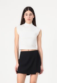Hvit kortermet topp uten ermer med høy krage, kombinert med ett svart innviklet skjørt. Glatt tekstur, minimalistisk design og tettsittende silhuett.