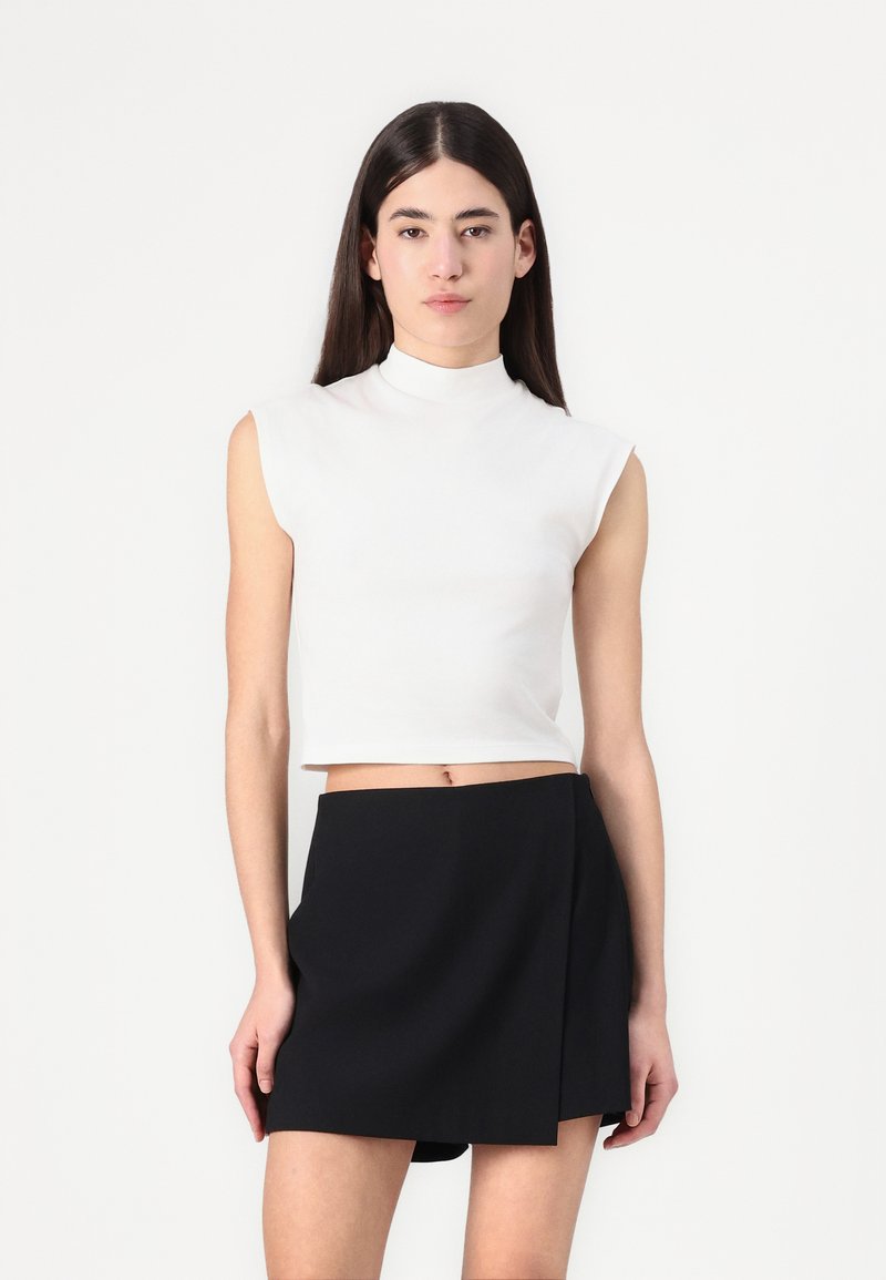 Hvit kortermet topp uten ermer med høy krage, kombinert med ett svart innviklet skjørt. Glatt tekstur, minimalistisk design og tettsittende silhuett.