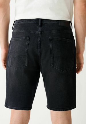 Henkilö, jolla on mustat farkkushortsit takataskuilla, seisoo yksivärisen vaalean taustan edessä, valkoinen paita osittain näkyvissä.