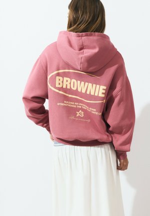 Woman wearing a pink hooded sweatshirt with "BROWNIE" and text printed on the back, paired with a long white skirt, facing away.