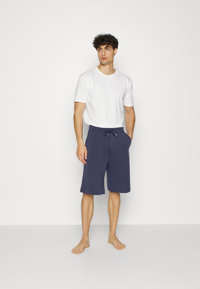 Bermudas azul-marinho com cintura elástica e cordão, combinadas com uma t-shirt branca simples. Modelo descalço em pé sobre um fundo branco.