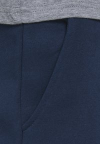 Pantaloni della tuta blu navy con una texture liscia, caratterizzati da una tasca laterale e un bordo di felpa grigia che sovrasta la parte superiore.