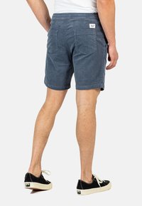 Bermudas de hombre en color marino hechas de pana con un corte relajado, que cuentan con un bolsillo trasero y una etiqueta con el logo en la cinturilla, combinadas con zapatillas negras.