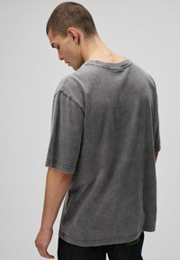 Młody mężczyzna z krótkimi włosami, ubrany w luźną, wyblakłą szaro-tshirt, stojący tyłem i patrzący w dół na jednolity jasny tle.