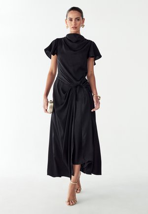 Black, knee-length dress with a draped neckline and knotted waist. Features short sleeves and flowing fabric, paired with gold strappy sandals.