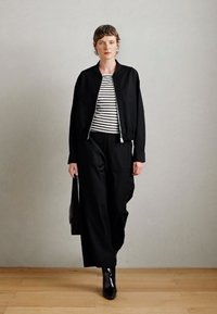 Veste bombardier noire sur une chemise rayée, associée à un pantalon noir ample. Le mannequin porte des bottines noires brillantes et tient un sac noir.