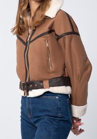 Veste en suédine marron avec un col en fausse fourrure crème, des accents en cuir, une poche zippée et une taille ceinturée. Manches droites.