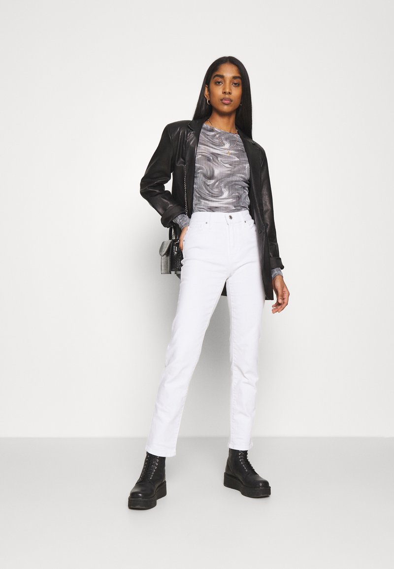 Blazer en cuir noir, haut gris à motifs, jean droit blanc et bottes noires à semelles épaisses. Accessoires minimaux incluant un petit sac.