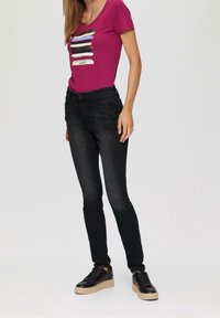 Mujer con una camiseta color frambuesa con diseño de pinceladas y texto "Sé consciente", jeans pitillo negros desgastados y zapatillas negras con suelas beige.