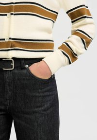 Osoba koja nosi bijeli kardigan s smešnim i crnim prugama, ruka u džepu crnih traperica s crnim pojasom.