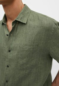 Koszula z krótkim rękawem na guziki, wykonana z ciemnozielonego lnu. Posiada pojedynczą kieszeń na piersi oraz pięć czarnych guzików z przodu, teksturowana powierzchnia.