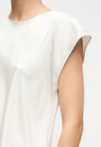 Λευκό αμάνικο τοπ με φαρδιά σχεδίαση ώμων, φορεμένο από ένα άτομο, που δείχνει τον άνω κορμό και το χέρι ενάντια σε απλό φόντο.