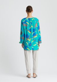 Blouse turquoise à manches longues et coupe ample avec des motifs abstraits en vert, bleu, marron et rose, associée à un pantalon beige clair et des sandales.