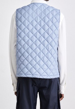 Personne portant une veste matelassée bleu clair par-dessus une chemise blanche à manches longues et un pantalon foncé, vue de dos sur un fond uni.