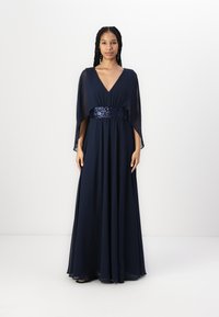Long navy blue gown with a deep V-neck and flowy cape sleeves. Sparkling sequined waist detail, smooth fabric, and an overall elegant shape.