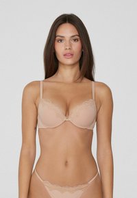 Žena s dlouhými tmavými vlasy, oblečená v béžové podprsence s krajkovým lemem a odpovídajícím spodním prádlem, stojící na pozadí jednoduchého světlého pozadí.