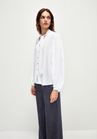 Weiße Bluse mit gefältelten langen Ärmeln, Schlüssel-Ausschnitt und goldenen Knöpfen. Kombiniert mit einer dunklen, gemusterten, weiten Hose.