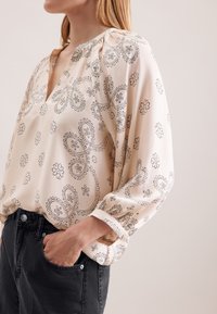 Lys beige bluse med sorte paisley- og blomster mønstre, med V-udskæring og lange ærmer, parret med mørke jeans. Blød stoftekstur.