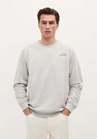 Sweatshirt gris clair avec des manches longues, un col rond côtelé et un texte brodé sur la poitrine gauche. Tissu doux en mélange de coton.