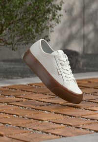 Baskets blanches texturées avec une semelle marron flottant au-dessus d'un sol en carreaux de bois, avec un arrière-plan flou de verdure et de mur en béton.