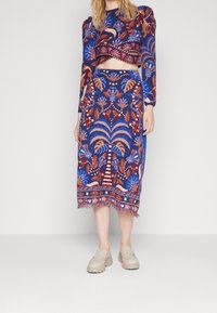Ensemble deux pièces bleu et rouge à motifs avec des manches bouffantes, un top court et une jupe longueur genoux avec ourlet à franges et motifs de palmiers.