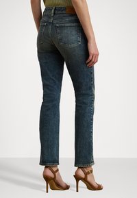 Högmidjade mörkblå jeans med rak passform, i en texturerad tyg, fem fickor och en läderpatch bak, kombinerade med bruna klackar.