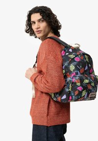 Batoh s černou tkaninou s barevnými geometrickými vzory, s nastavitelnými popruhy a přední kapsou. Nese ho osoba s kudrnatými vlasy.