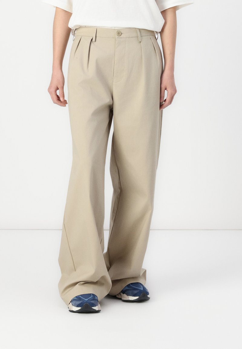 Pantalons beige à jambes larges avec plis à l'avant, fabriqués en tissu léger. Assortis à des chaussures de sport bleu et blanc, le tout sur fond uni.