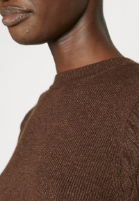 Maglione lavorato a maglia marrone con colletto a coste e motivo testurizzato, mostrato con la spalla e il collo del modello visibili.