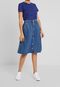 Blue patterned T-shirt with short sleeves paired with a button-down denim skirt. Skirt features an elastic waistband and side pockets. White sneakers.