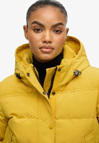 Gul puffjakke med lynlås foran, stor hætte og sorte hardwaredetaljer. Glat, polstret tekstur og syede detaljer.