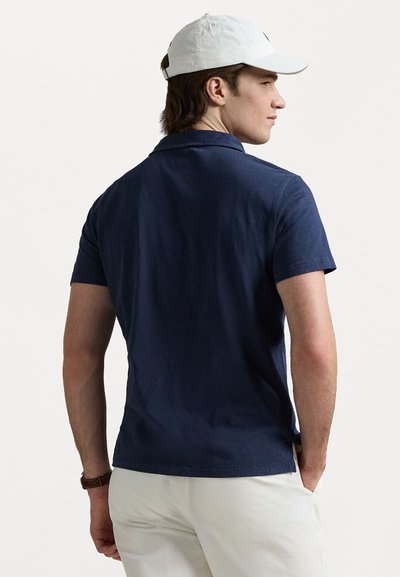 Polo shirt bleu marine à manches courtes en mélange de coton, avec un col classique et un ourlet droit. Porté avec un pantalon blanc.