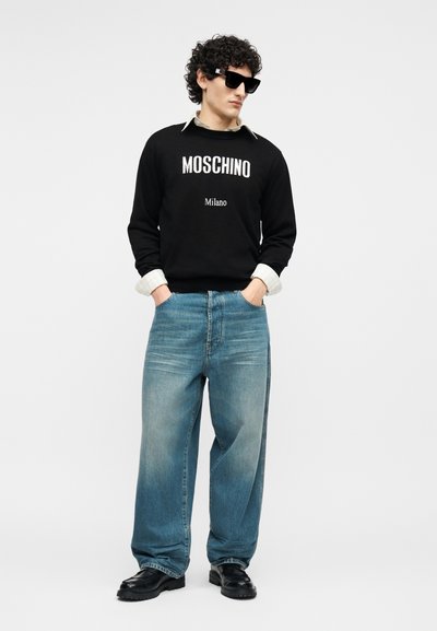 Homme aux cheveux bouclés portant un sweat-shirt noir Moschino, un jean fluide bleu clair, des chaussures noires et des lunettes de soleil noires, posant les mains dans les poches.