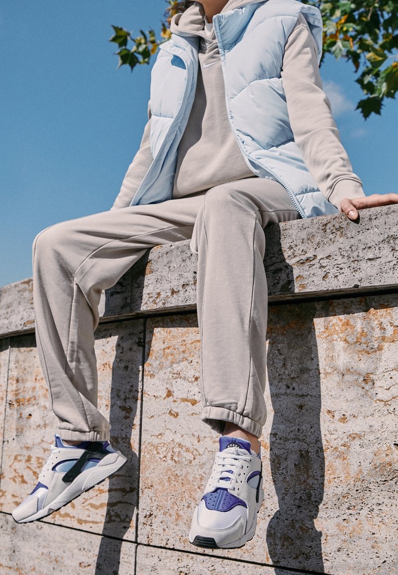 Veste matelassée bleu clair sur un sweat à capuche gris, associée à un pantalon de jogging gris et des baskets blanches avec des accents violets ; assis sur une surface en pierre.