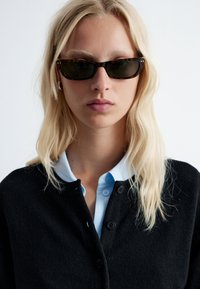 Schwarze Strickjacke mit Knöpfen, getragen über einem hellblauen Hemd mit Kragen. Das Model trägt dunkle, schmale Sonnenbrillen mit einem Schildpattgestell.