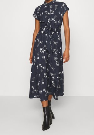 Kobieta w granatowej, kwiatowej sukience midi z krótkimi rękawami, z wiązaniem z przodu w talii i czarnych botkach, stojąca na jednolitym tle.