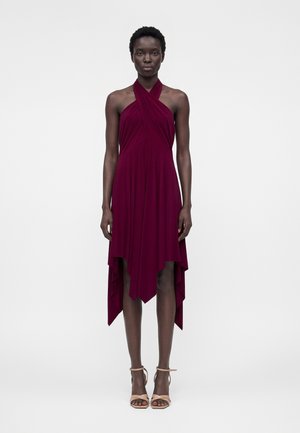 Mujer de pie mirando hacia adelante, vestida con un vestido sin mangas color burdeos con escote halter, dobladillo asimétrico y sandalias beige de tacón alto.