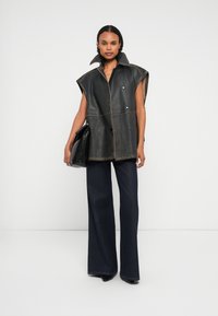 Donna che indossa un gilet in pelle nera senza maniche con colletto alto, jeans scuri a gamba larga, scarpe nere e tiene una grande borsa a tracolla nera.