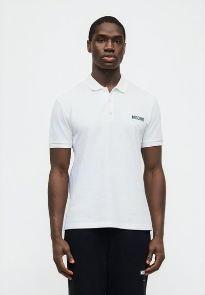 Άνδρας που φοράει λευκό πόλο Moschino και μαύρο παντελόνι, στέκεται μπροστά σε απλό λευκό φόντο, κοιτώντας ευθεία με ουδέτερη έκφραση.