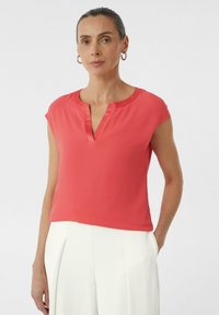 Koraliowa bluzka z krótkimi rękawami, dekoltem w kształcie litery V i gładką fakturą. W zestawieniu z białymi spodniami, które mają fałdy, co nadaje elegancki wygląd.