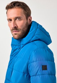 Giacca puffo blu con cappuccio, caratterizzata da cuciture trapuntate, texture liscia e un'appliqué logo nero sulla spalla sinistra.