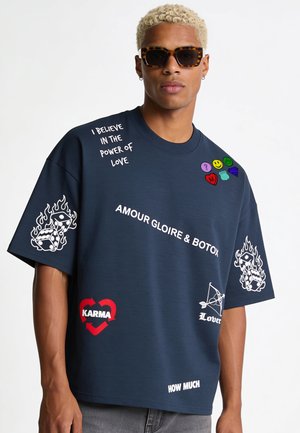 Man met zonnebril en een donkerblauw oversized T-shirt met witte en gekleurde grafische tekst en pictogrammen waaronder dobbelstenen, hart en uitdrukkingen.