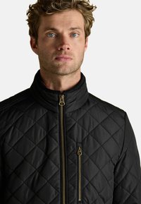 Mężczyzna z niebieskimi oczami i krótkimi, kręconymi blond włosami, noszący czarną pikowaną kurtkę z widocznymi kieszeniami na zamek, zwrócony przodem.