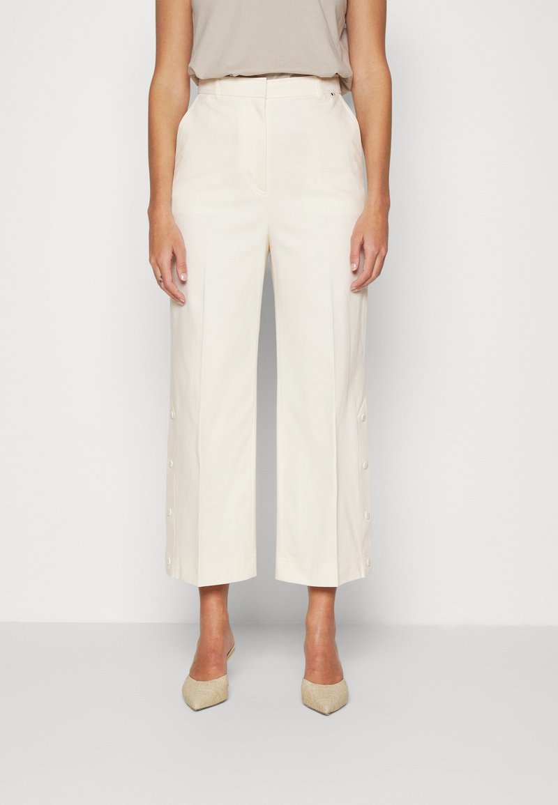 Femme portant un pantalon crème taille haute, jambes larges avec boutons sur le côté et des talons pointus beige, debout devant un fond clair uni.