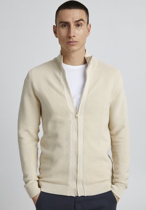 Beige zip-up genser med ribbet tekstur og høy krage. Har en glatt glidelås og tettsittende mansjetter. Brukt over en hvit t-skjorte.