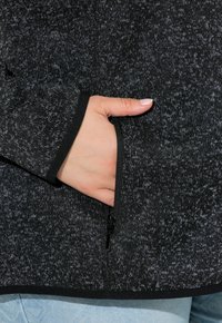 Giacca in pile nera e grigia con una tasca con zip e bordi neri a contrasto. Il materiale è morbido e ha un aspetto lavorato a maglia.
