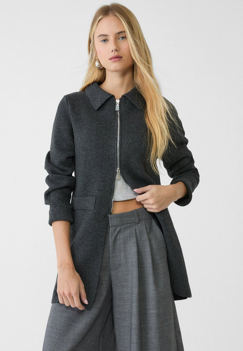 Vêtements d'extérieur tricotés gris foncé avec un col, fermeture éclair sur le devant et poches. Présente des manches retroussées et une coupe ajustée évasée.