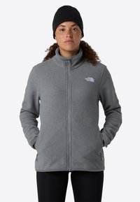 Donna con capelli ricci che indossa un berretto nero e una giacca in pile grigia con zip, con le mani nelle tasche, in piedi davanti a uno sfondo neutro.