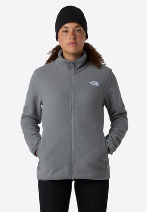 Donna con capelli ricci che indossa un berretto nero e una giacca in pile grigia con zip, con le mani nelle tasche, in piedi davanti a uno sfondo neutro.