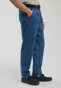 Mėlynos dryžuotos kelnės laisva forma, su raukšlėta priekiu ir šoninėmis kišenėmis, derinamos su tamsiai mėlynais kasdieniais batais.